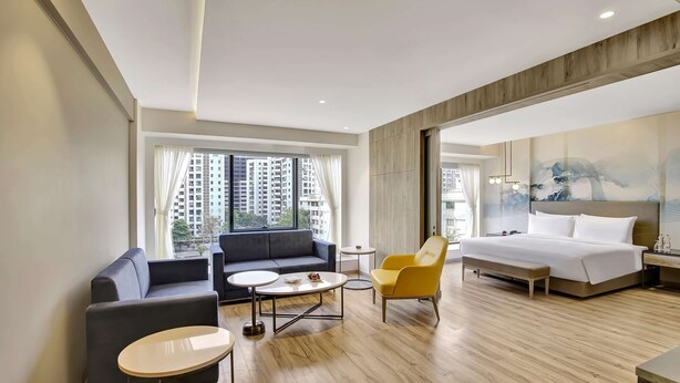 Habitación de hotel moderna con sofá, un sillón amarillo, una mesa de centro redonda, cama y un ventanal con cortinas.