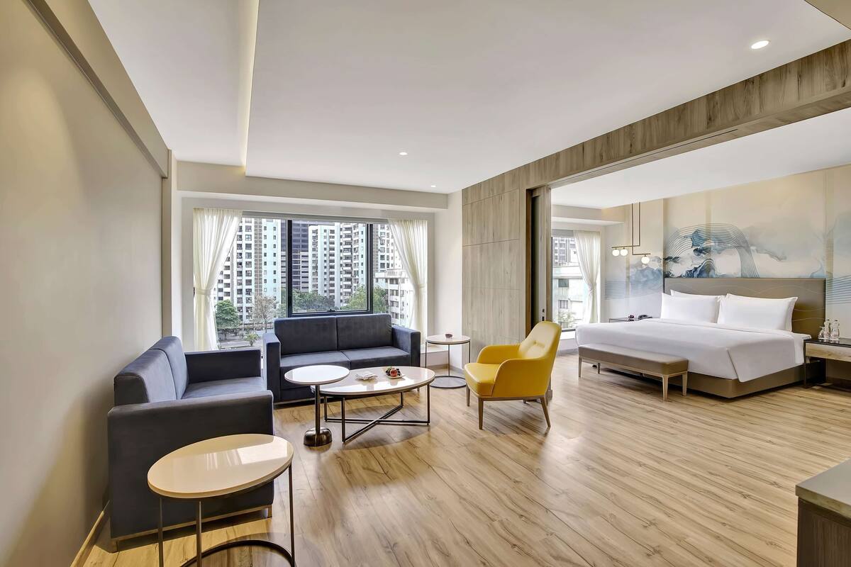 A modern hotel room with a sofa, a yellow armchair, a round coffee table, a bed, and a large window with curtains.