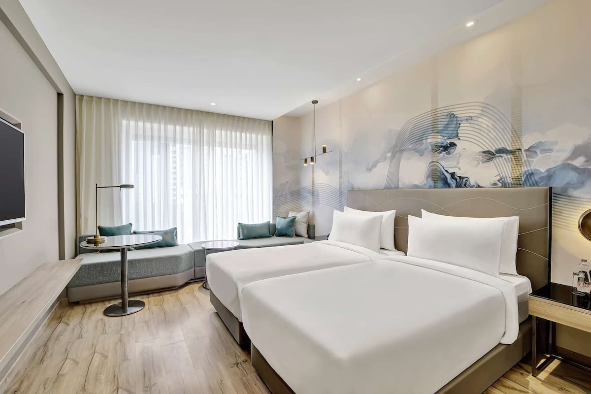 A modern hotel room with a large bed, a sofa bed, a small table, and a wall-mounted TV.