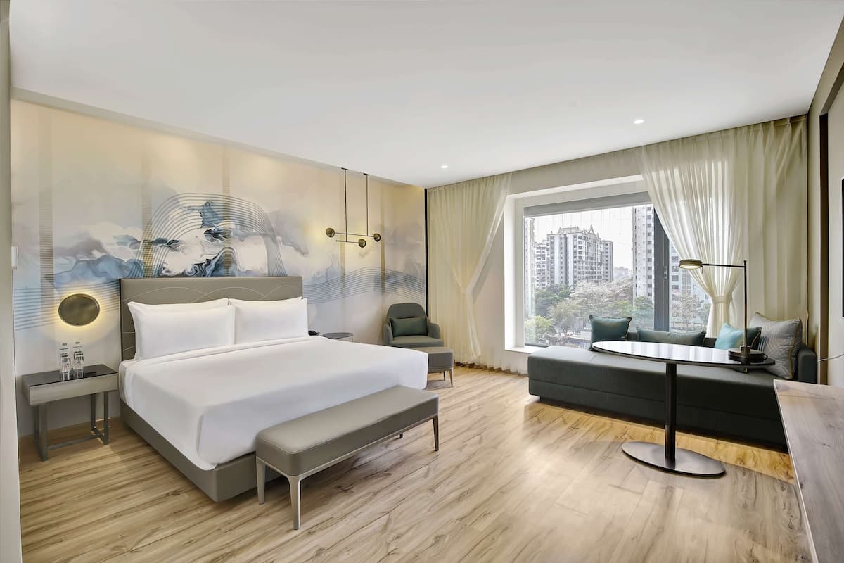A modern hotel room with a large bed, a seating area with a table, a view of the city through large windows, and a wall with a decorative mural.