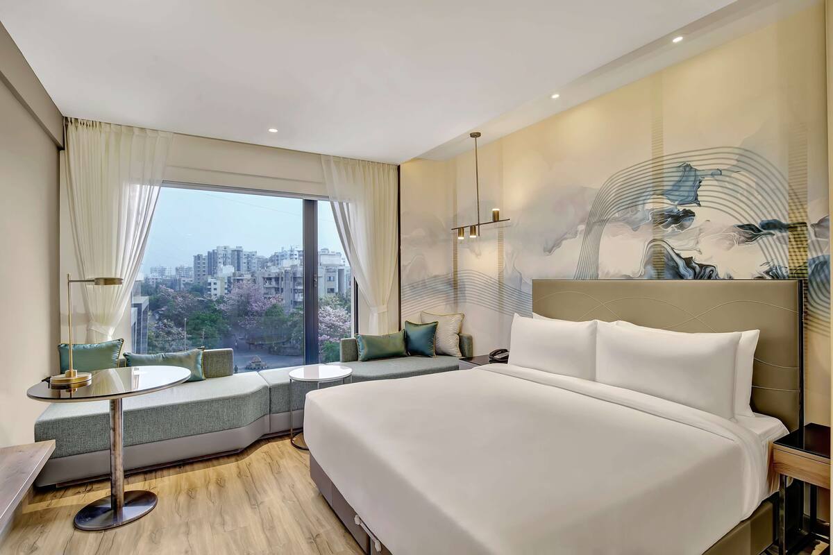 A modern hotel room with a large bed, a seating area with a view, and a cityscape visible through the window.