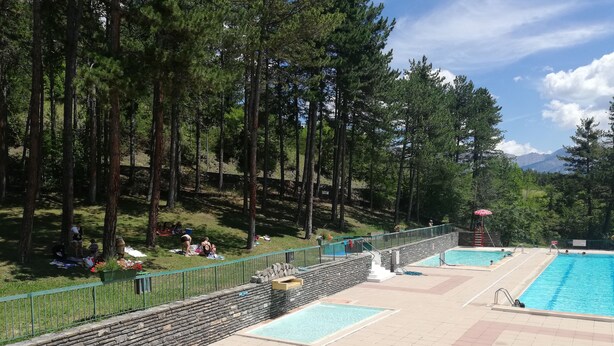Un área de piscina con pared de piedra y suelo de baldosas, rodeada por una valla y árboles.