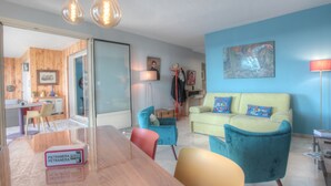 Ein modernes Wohnzimmer mit einem Holzesstisch, smaragdgrünen und gelben Stühlen, einem hellgrünen Sofa mit blauen Kissen und einem Gemälde an der Wand.