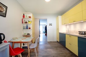 A compact kitchen with a dining area, featuring a wooden table, red chairs, and a white shelf with various kitchen items.