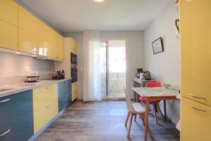A compact kitchen with yellow and blue cabinets, a dining table with chairs, and a view of the outside through a glass door.