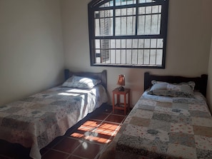 A bedroom with two beds, a window with blinds, a small table with a lamp, and a patterned rug. - Spacious house for family (Guarujá)