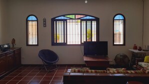 Un salon équipé d’un téléviseur, d’un ventilateur et d’un canapé. Il y a deux fenêtres avec des barreaux et un ventilateur de plafond.