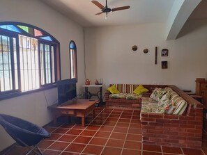 A room with a tiled floor, a wooden desk, a chair, a sofa with cushions, a ceiling fan, and a window with bars. - Spacious house for family (Guarujá)