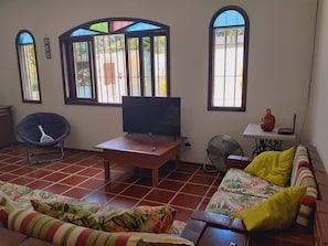 A living room with a TV on a wooden stand, a sofa with floral cushions, a chair, and a fan. - Spacious house for family (Guarujá)