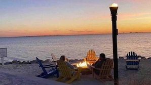 Eine Strandansicht mit zwei Personen, die in Adirondack-Stühlen einem Feuerkorb gegenüber sitzen.