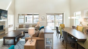 A spacious living room with a sofa, coffee table, and dining table. There is a TV mounted on the wall and a ceiling fan. Large windows provide natural light and a view of the outside.