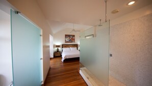 Een hotelkamer met een houten vloer, een bed met witte lakens, een nachtkastje met een lamp en een plafondventilator.