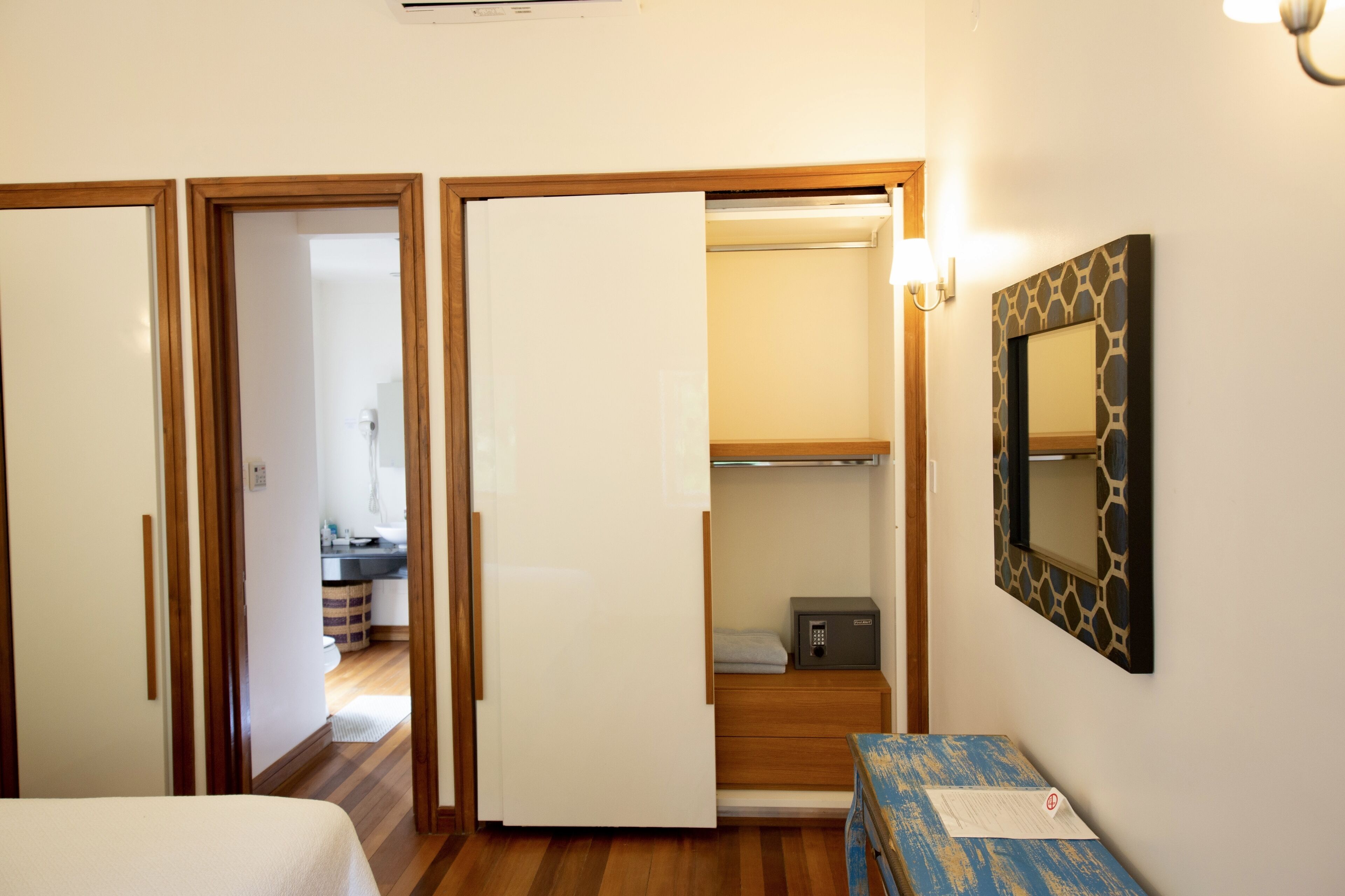 A hotel room with a wooden floor, a bed, a desk, a wardrobe, and a bathroom visible through an open door.