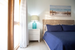 Quarto com cama, mesa de cabeceira com abajur, quadro de uma praia e janela com cortinas.