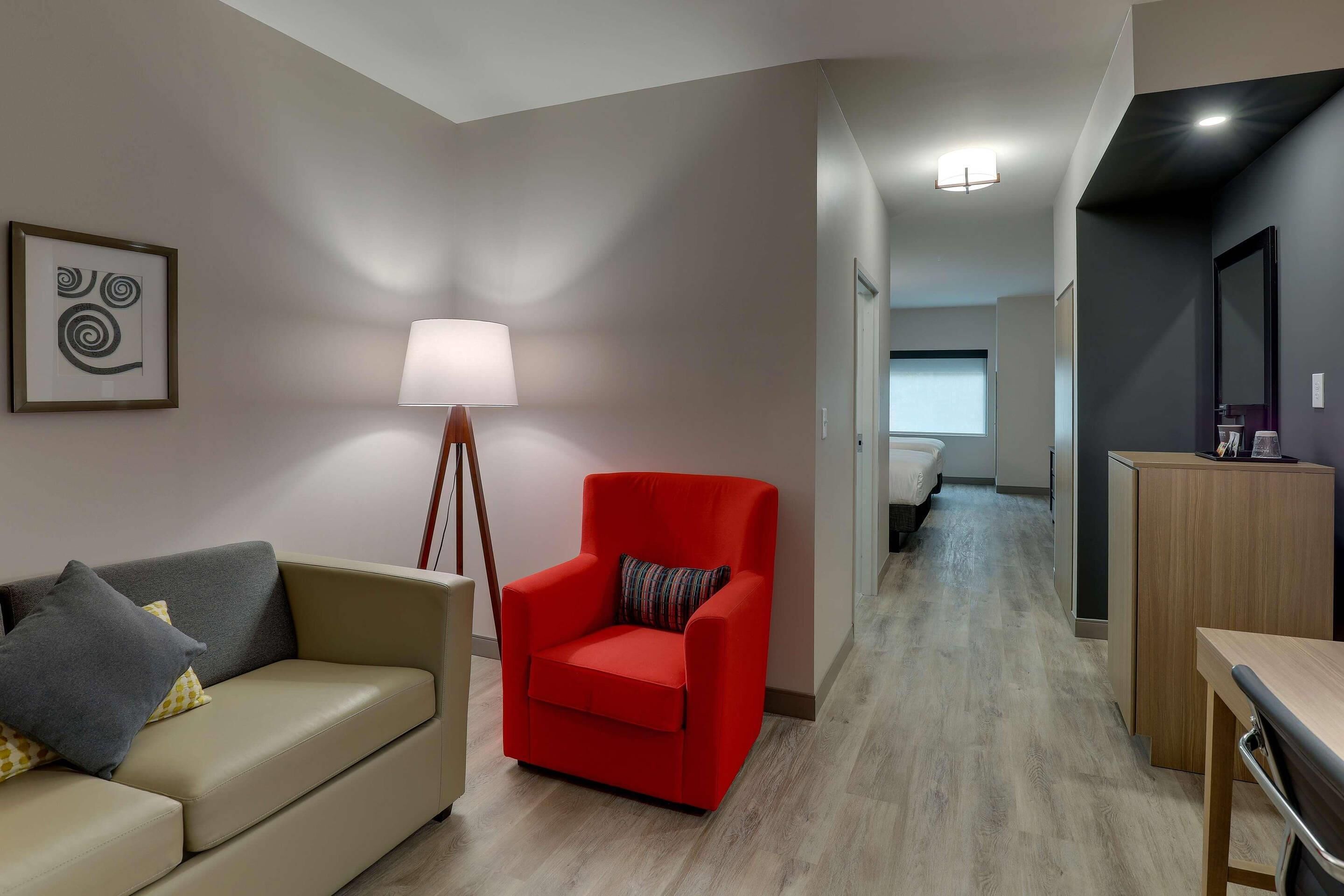 A modern hotel room with a sofa, a red armchair, a lamp, a desk, and a mirror.