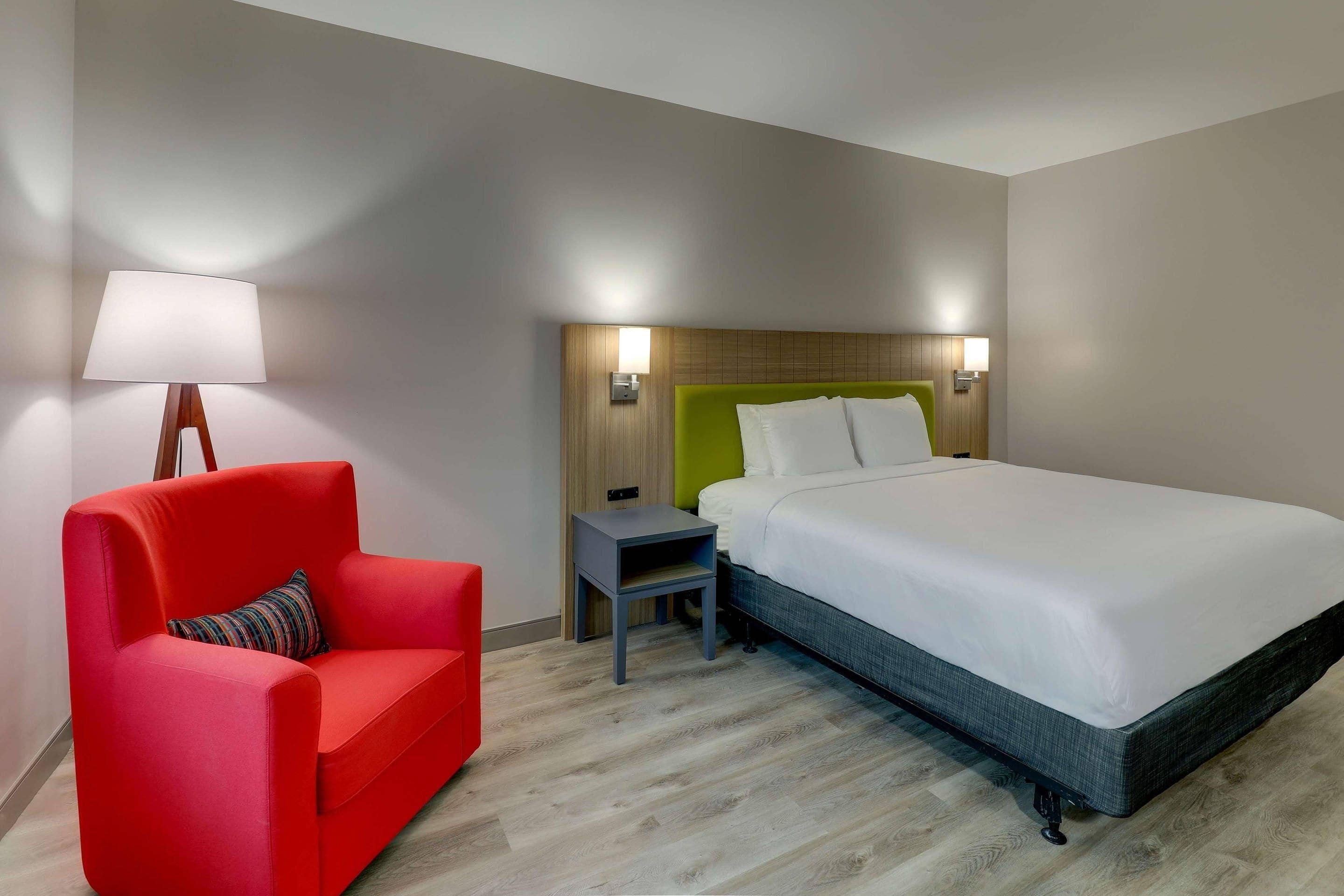 A hotel room with a bed, a red chair, a nightstand, and a lamp.