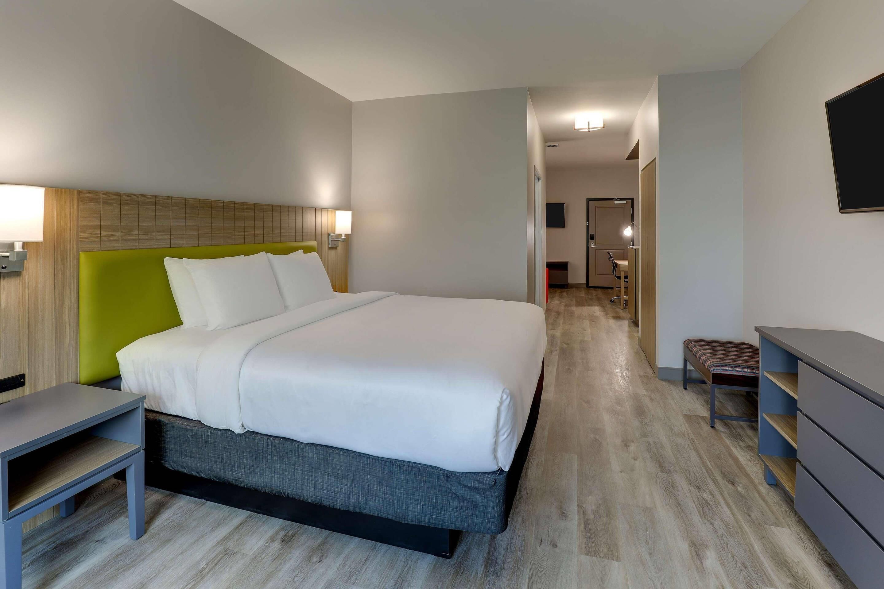 A modern hotel room with a bed, bedside table, bench, and a desk.