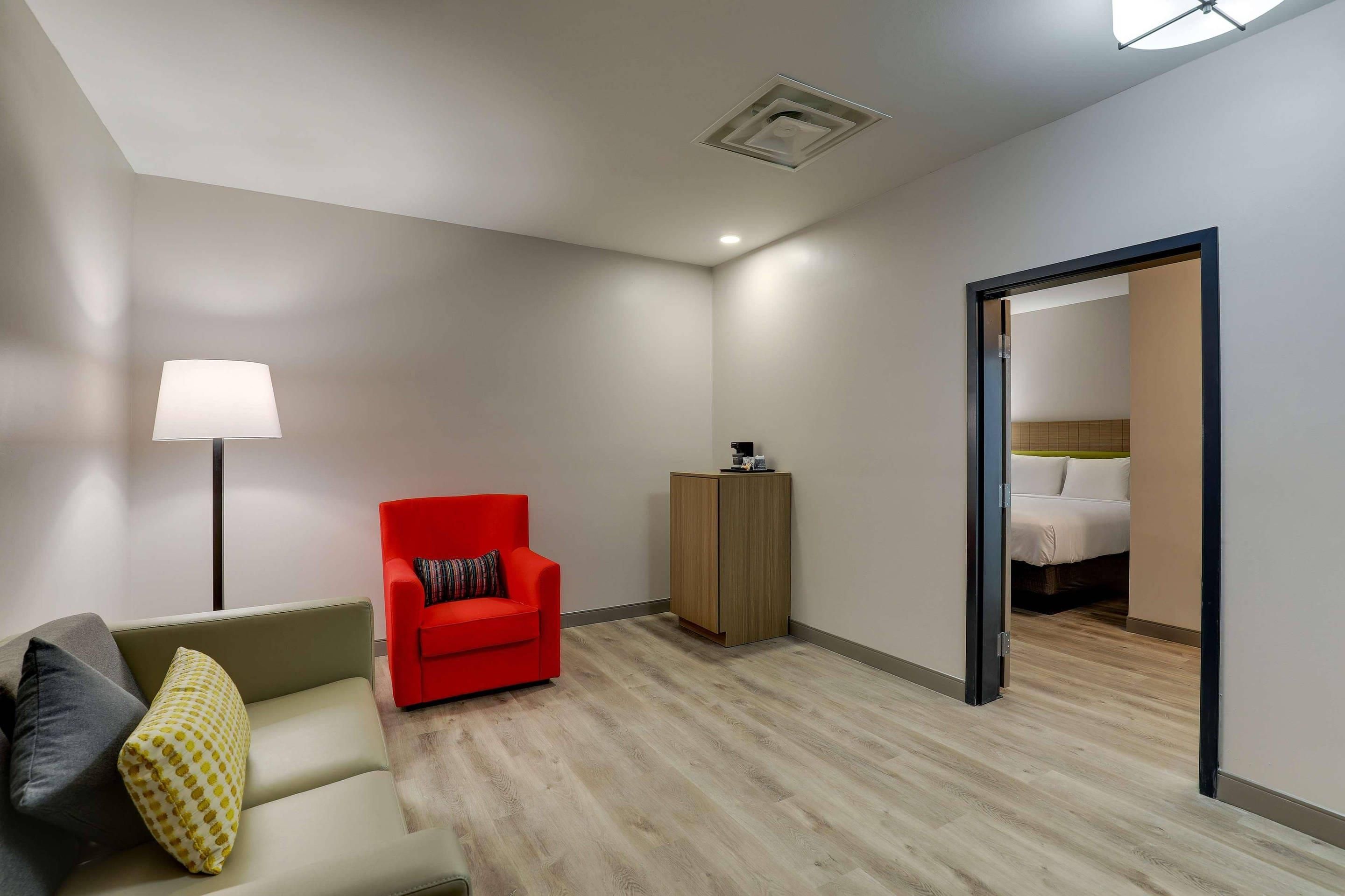 Quarto de hotel moderno com sofá, uma poltrona vermelha, um armário de madeira e uma cama com cabeceira branca.