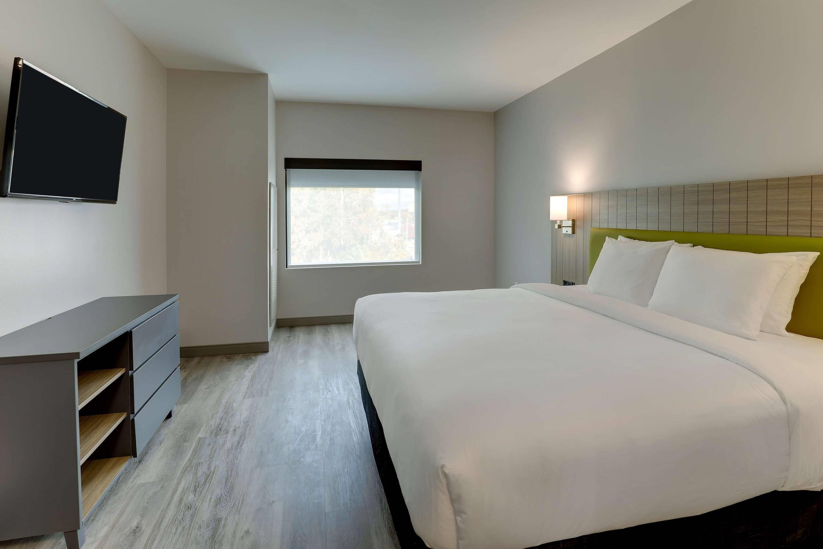 Habitación de hotel con cama, escritorio, televisor, ventana y lámpara.
