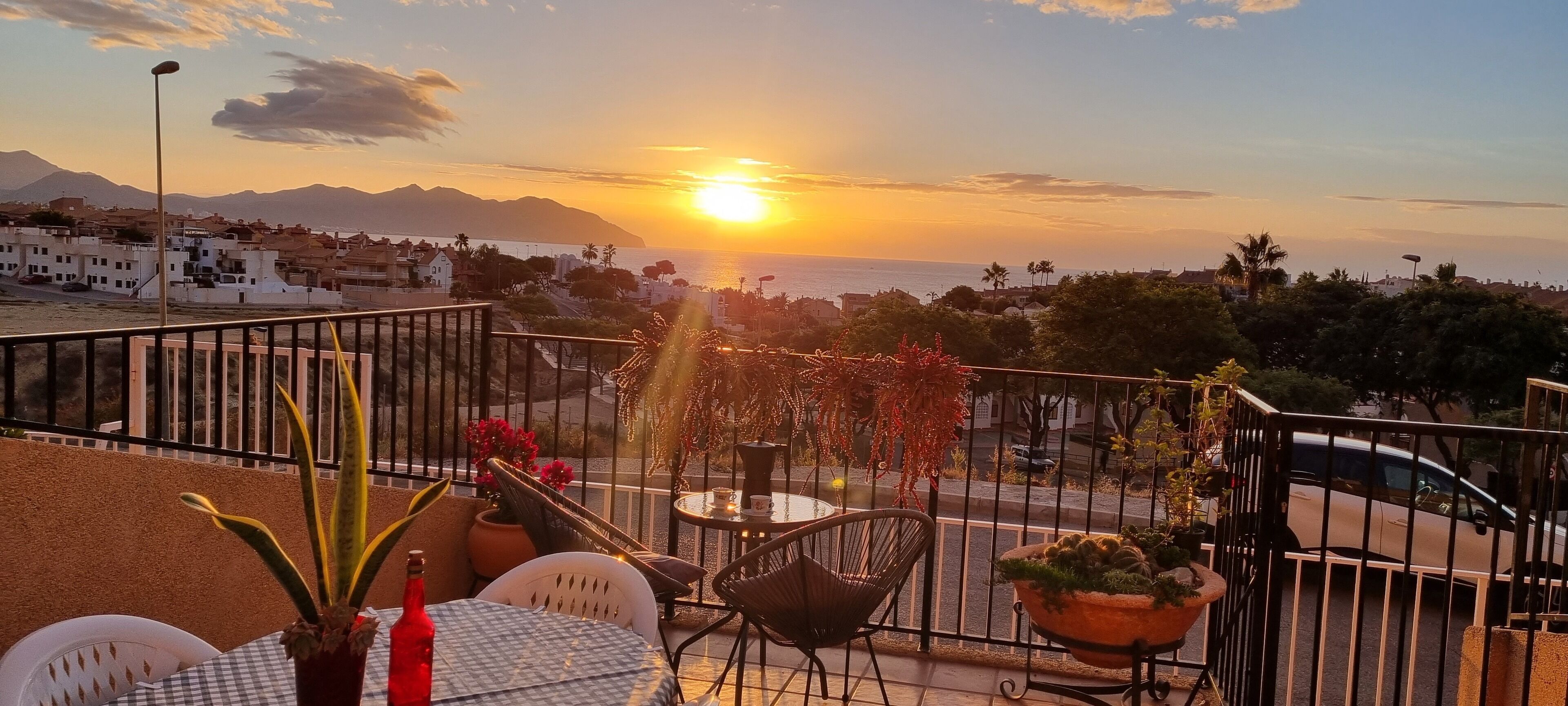 A balcony with a table and chairs, overlooking a sunset over the sea with buildings in the background.