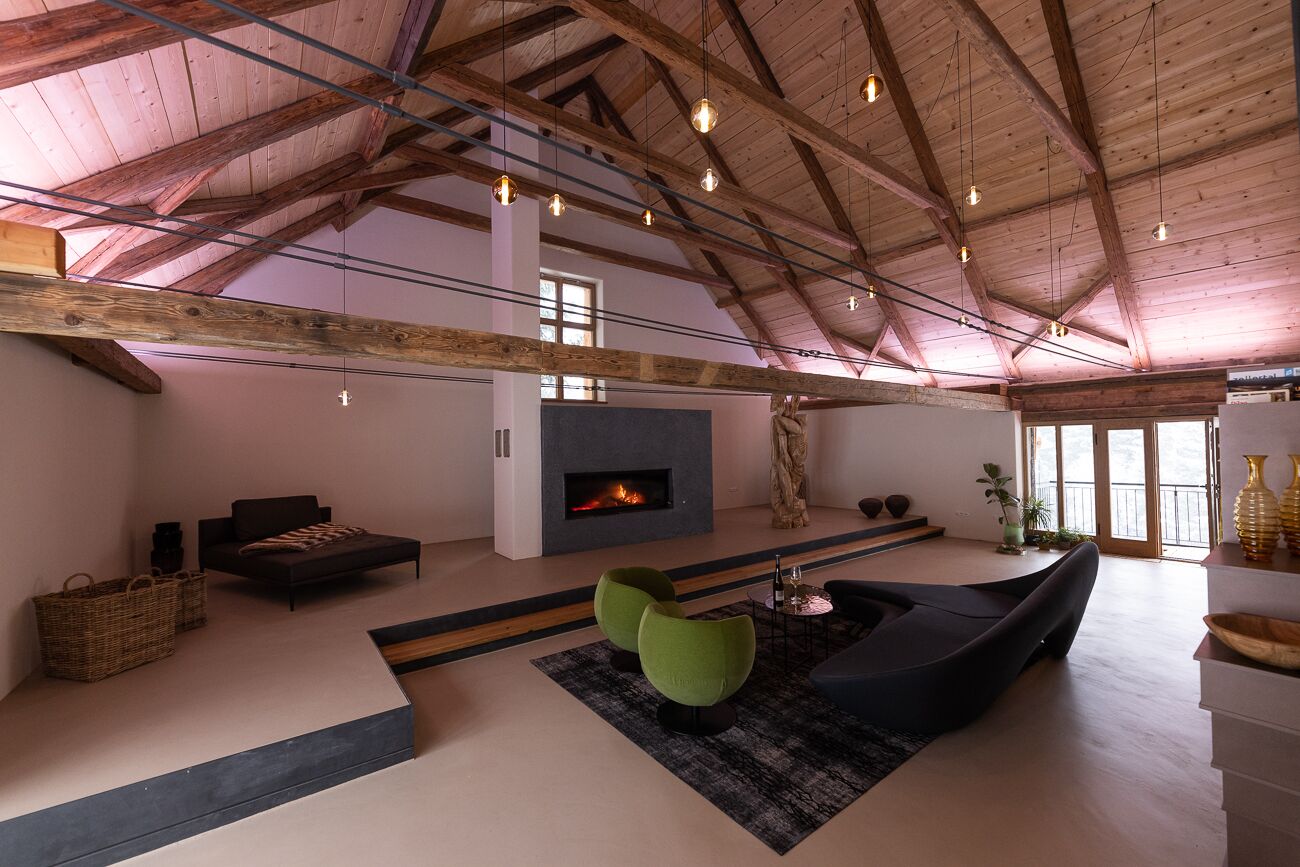 Ein geräumiges Wohnzimmer mit hoher Decke, freiliegenden Holzbalken, Kamin, moderner Sitzgelegenheit und großen Fenstern.