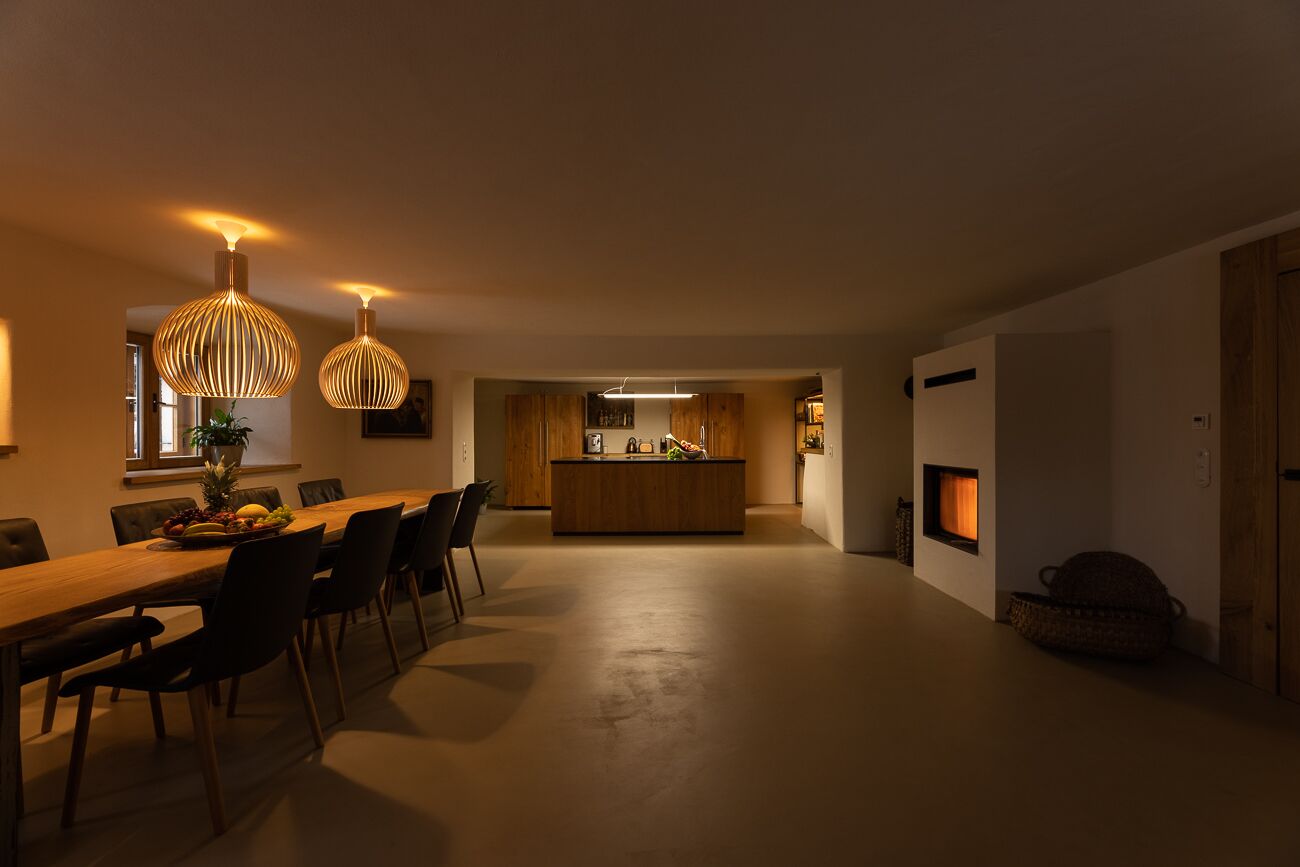 A modern dining area with a long wooden table and chairs, a kitchen in the background, and pendant lighting.