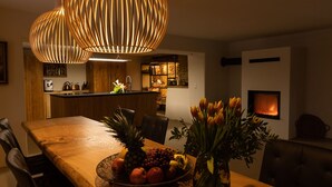Un comedor con una mesa de madera, sillas, un frutero, un jarrón con flores y una lámpara colgante moderna.