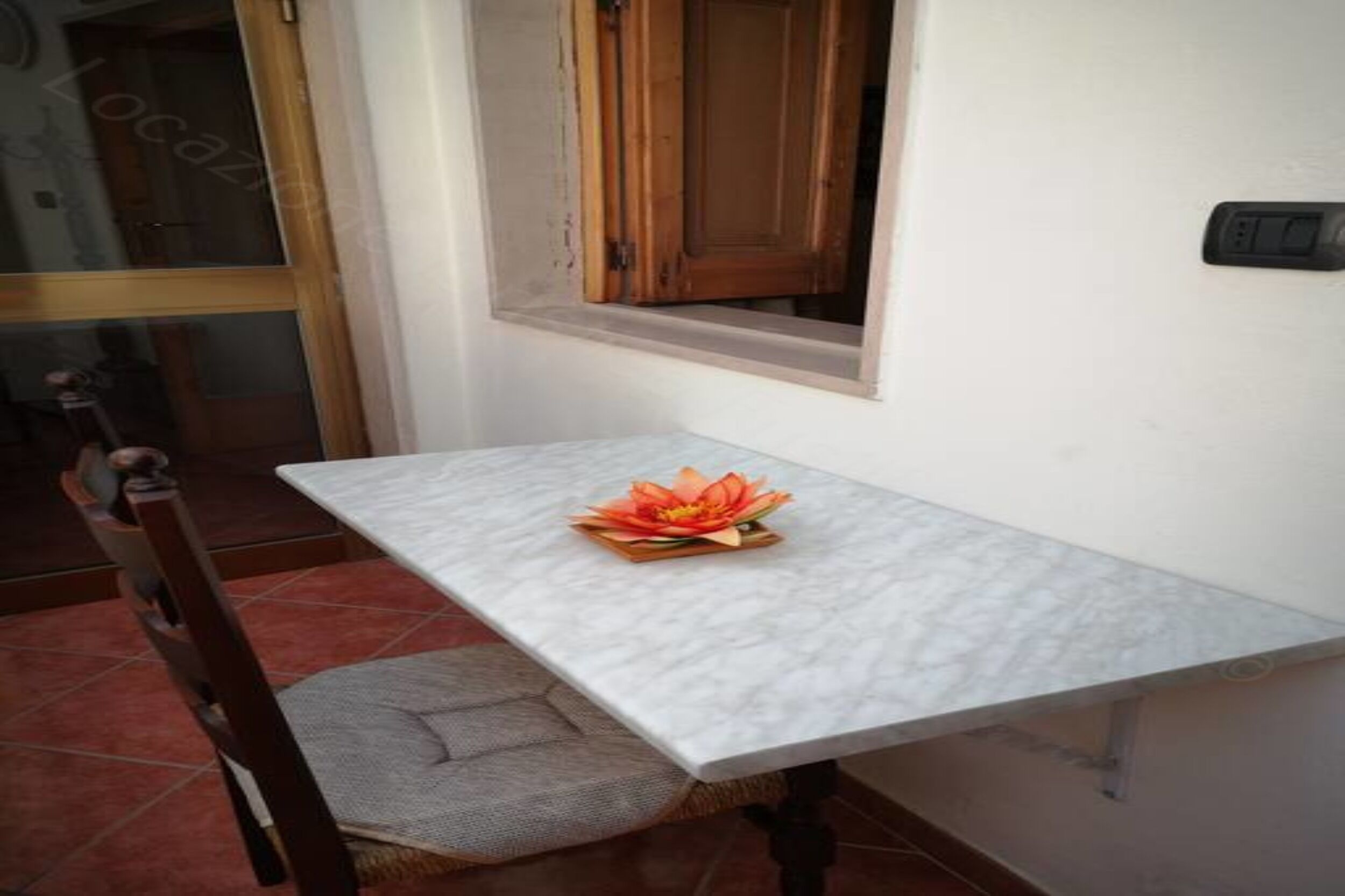 Une petite table agrémentée d’un arrangement floral décoratif, une chaise en bois et une fenêtre avec un volet en bois.