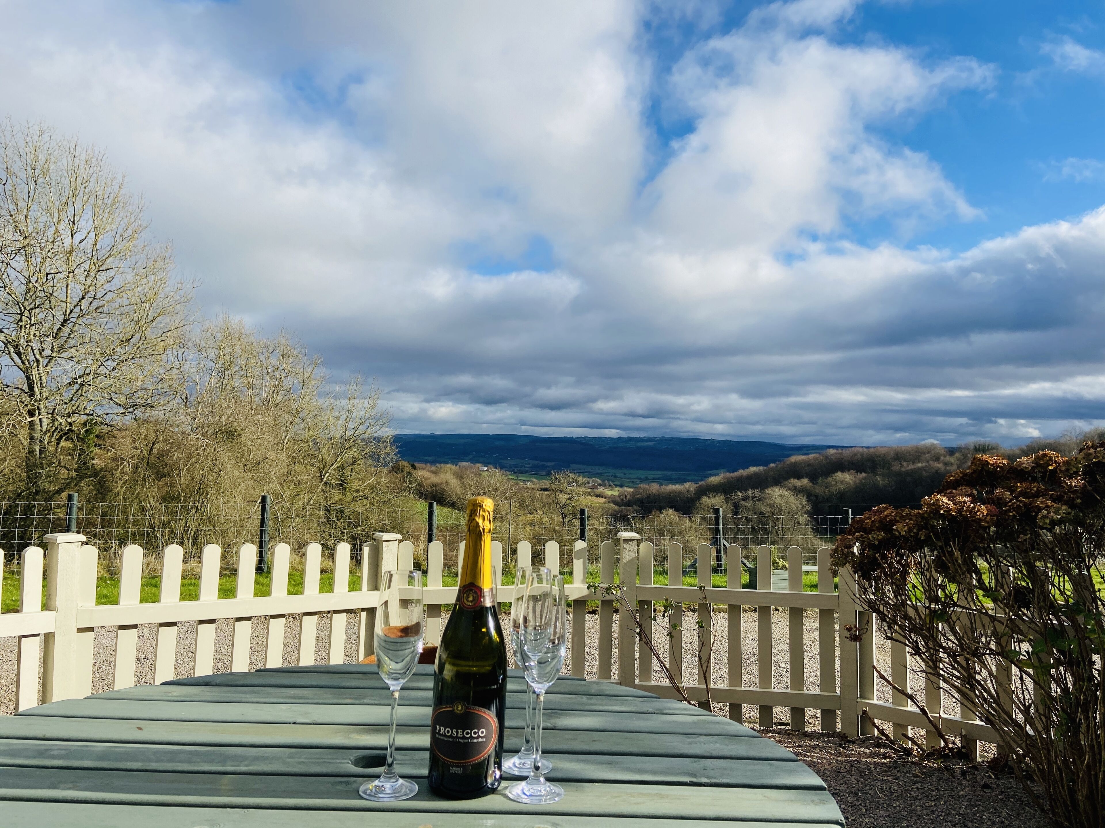 Una mesa con una botella de Prosecco y dos copas sobre una terraza de madera, con vistas a un paisaje pintoresco con una cerca y árboles.