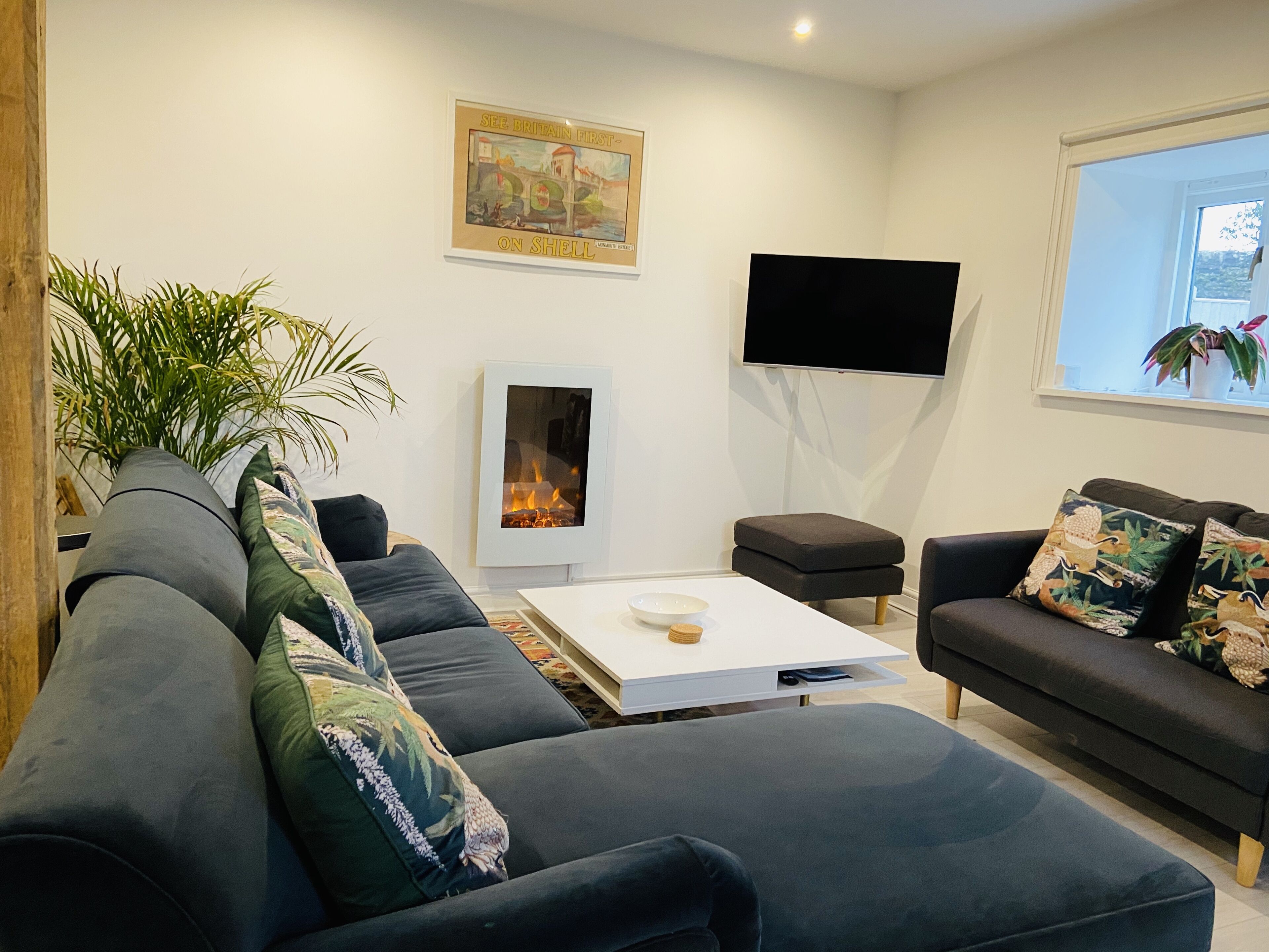 A modern living room with a large grey sofa, a white coffee table, a flat-screen TV mounted on the wall, and a fireplace.