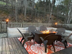 An outdoor patio with a fire pit table, four chairs, and a wooden deck.