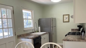 Una cocina con una mesa de madera, sillas blancas, un refrigerador, un lavavajillas y una ventana con vista a árboles.