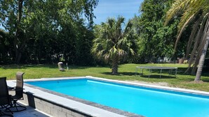 A rectangular swimming pool with a tiled deck area, surrounded by greenery and trees.