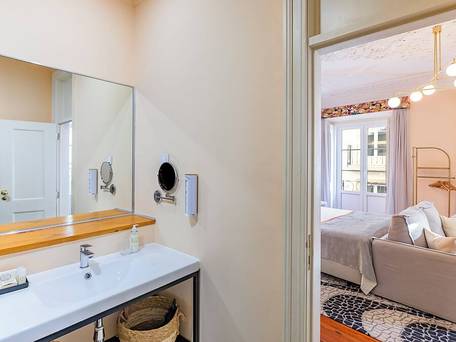 Un baño con un espejo grande, lavamanos y barra para toallas. Hay una puerta que da a otra habitación.