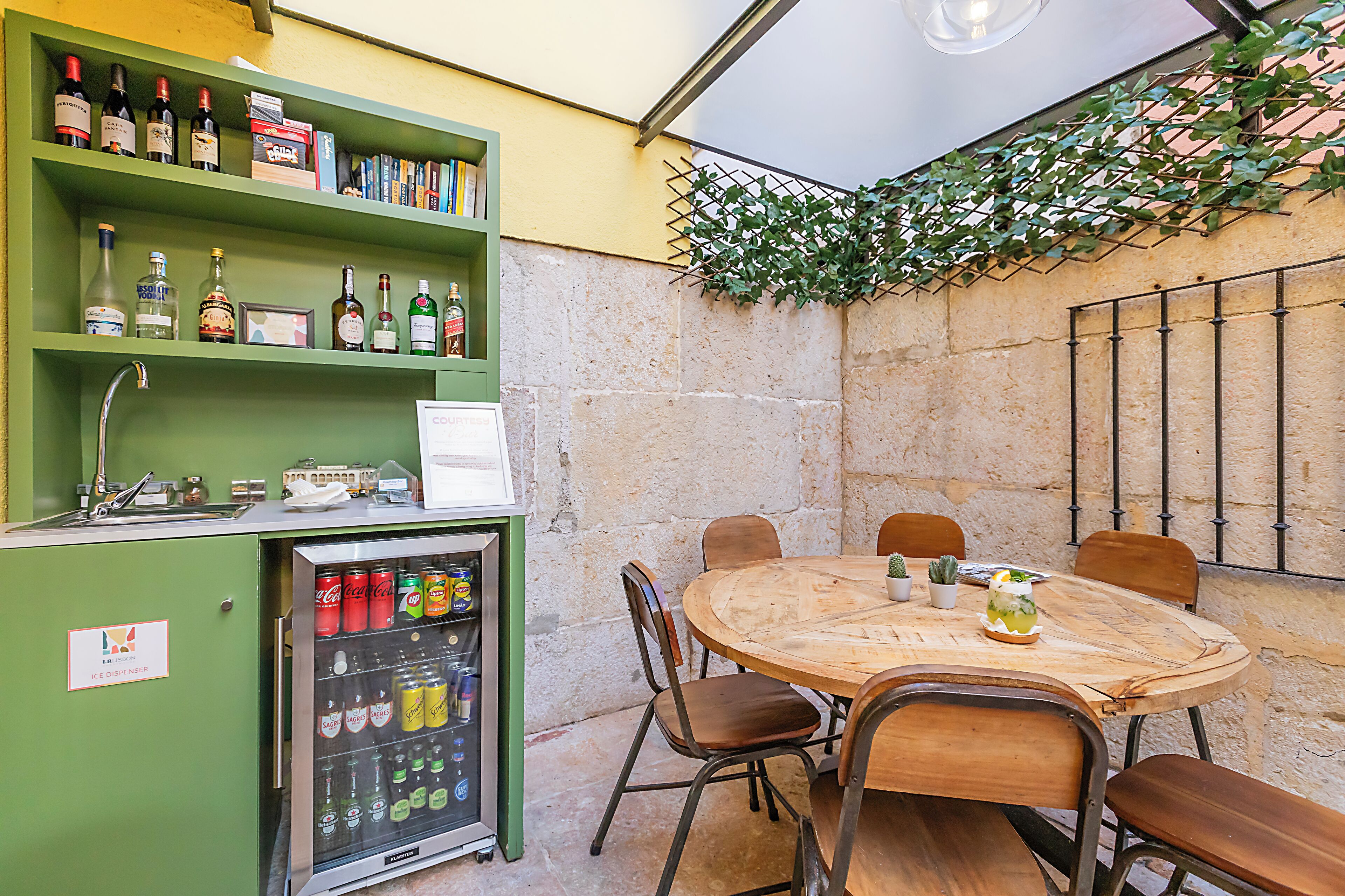 A small, enclosed outdoor dining area with a wooden table and chairs, a vending machine with beverages, and a green shelving unit with various bottles.