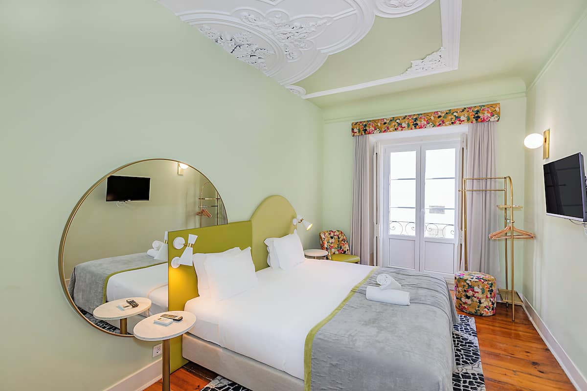 A hotel room with a bed, a round mirror, a small table, a chair, a television, and a window with curtains.
