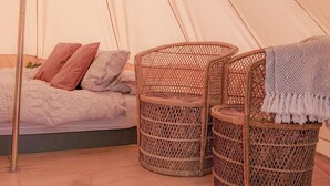 Interior de una tienda de campaña con una cama, dos sillones de mimbre y una manta.