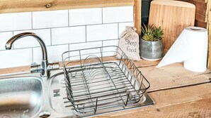 Una cocina con pared de madera, pileta de metal, escurreplatos de alambre, un bol de vidrio, un bol de cerámica, una decoración de gallo, una planta en maceta y un rollo de toallas de papel.