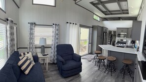 A modern living room with a sofa, armchair, and a table with a lamp. There is a kitchen area with a stove and bar stools.