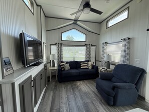 A compact living space with a ceiling fan, a flat-screen TV, a dark-colored sofa, a chair, and a window with patterned curtains. - Cozy Cottage Park Model at Upscale RV Resort in South Haven, Michigan. (South Haven)