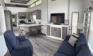 A modern kitchen with a central island, bar stools, a seating area with a sofa, and a television. - Cozy Cottage Park Model at Upscale RV Resort in South Haven, Michigan. (South Haven)
