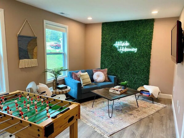 A cozy living room with a foosball table, a blue sofa, and a green wall with a neon sign. - Hillside Hideaway Boho 3BR Home w Gameroom, Waffle Bar, Gas Fire Pit, Mtn Views! (Canton)