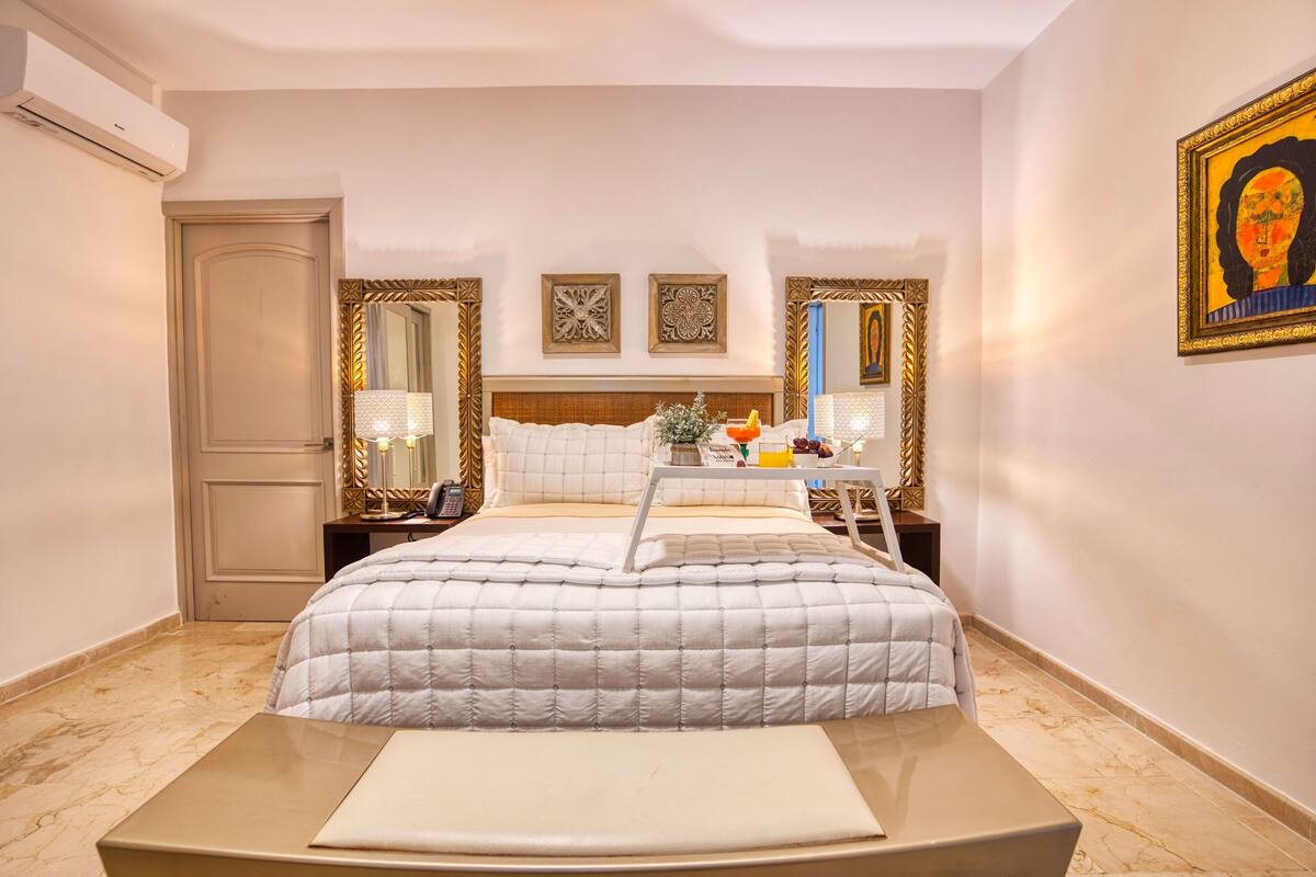 A hotel room with a bed, a bedside table, a nightstand, a mirror, and a wall-mounted artwork.