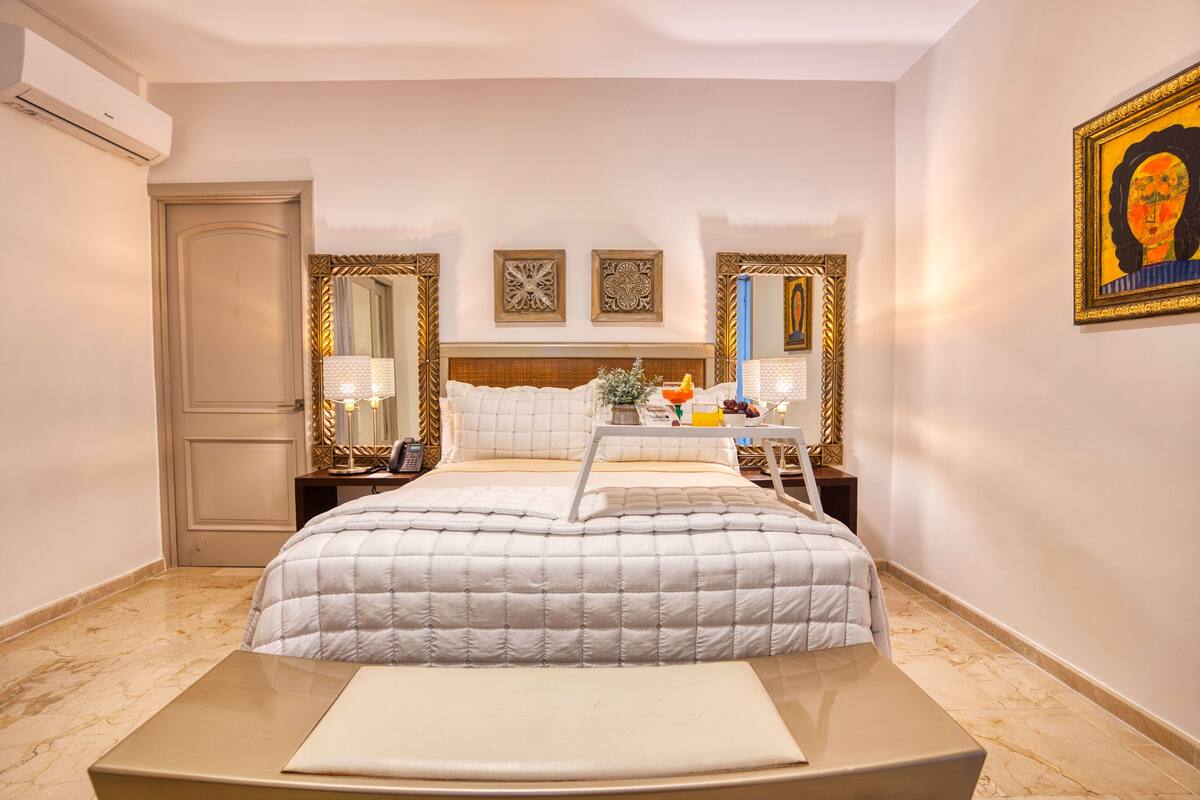 A hotel room with a large bed, a bedside table with a lamp, a tray with a glass of orange juice, and a framed picture on the wall.
