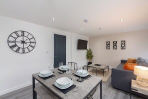 A modern living room with a dining area, a grey sofa, a TV mounted on the wall, and a large wall clock.