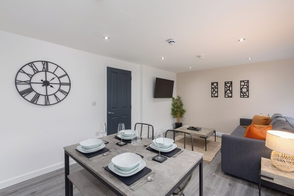 Un salón moderno con comedor, un sofá gris, un televisor montado en la pared y un reloj de pared grande.