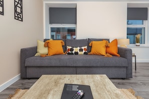 A living room with a grey sofa, orange cushions, a wooden coffee table, and a remote control on it.