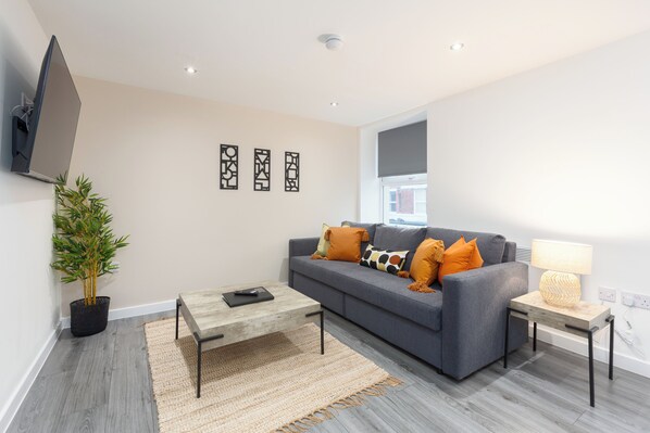A modern living room with a grey sofa, a wooden coffee table, a small side table with a lamp, a potted plant, and abstract wall art.