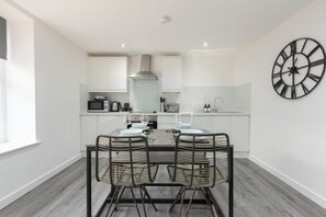 A modern kitchen with white cabinets, a stainless steel range hood, and a dining area with two wicker chairs and a wooden table.