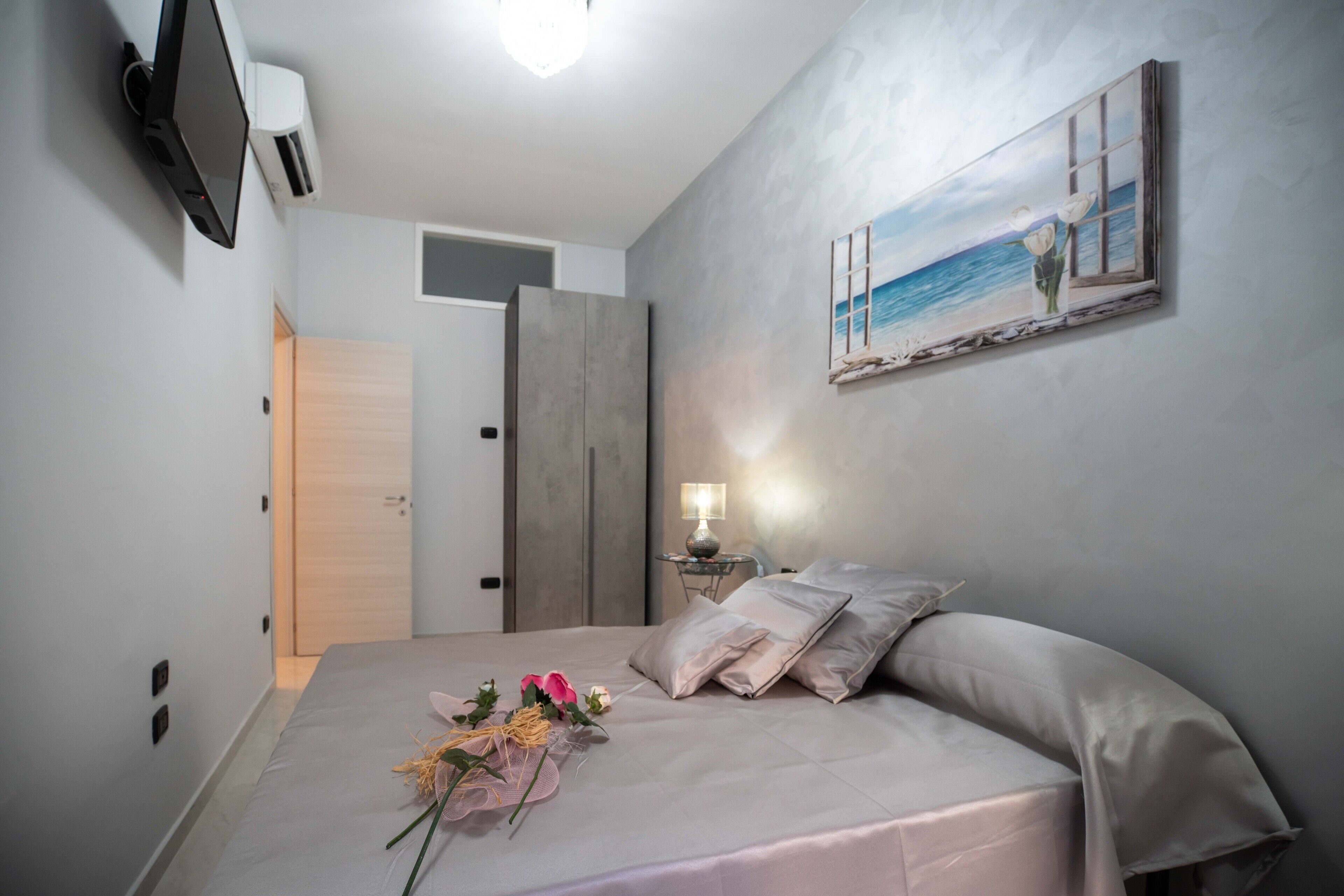 A bedroom with a bed, a painting of a beach scene, a bedside table with a lamp, and a wall-mounted TV.